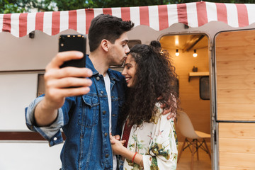 Sticker - Image of man kissing pretty woman and taking selfie on cellphone near house on wheels outdoors