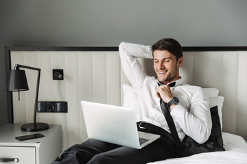 Image of happy young man lying on bed with laptop computer in hotel room during business trip