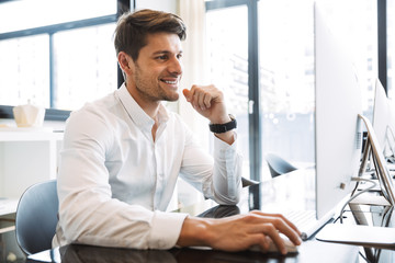 Poster - Image of happy businesslike man sitting at table and working on computer in office