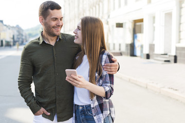 Smiling young woman holding smartphone and looking at her boyfriend on street