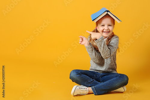 Fotografía Cheerful attractive little student girl is sitting on the floor with a book on her head and pointing to the side