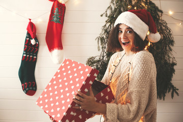 Stylish surprised girl in santa hat opening gift box in christmas decorated room. Young woman in festive cozy sweater opening present on background of modern christmas tree, lights and stockings