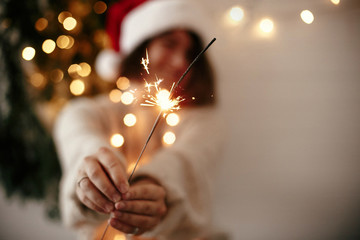 Wall Mural - Happy New Year eve party atmosphere. Sparkler burning in hand of stylish girl in santa hat on background of modern christmas tree light in dark room. Woman with fireworks. Happy Holidays