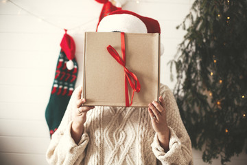 Wall Mural - Stylish happy girl in santa hat holding christmas gift box in front of face on background of modern christmas tree, lights and stockings. Young hipster woman in cozy sweater holding present