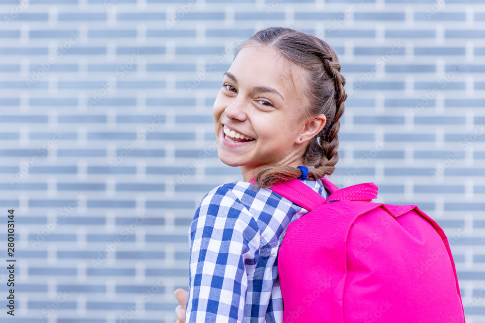 Beautiful student teenager schoolchild with backpack looking at camera. Smiling cute child with bag. Teen girl with braided hair against a brick wall outdoors. Childhood and Back to school concept. - obrazy, fototapety, plakaty 