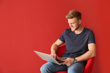Handsome Young Man With Laptop Sitting On Chair Against Color Background