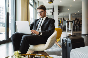 Poster - Portrait of caucasian young businessman sitting on armchair with laptop computer and suitcase in hotel hall