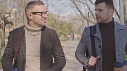 Poster - Two stylish adult men with cups of coffee speaking while walking in the park