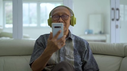 Wall Mural - Happy senior man listening music on a smartphone with headphones while sitting on the sofa in living room at home. Shot in 4k resolution