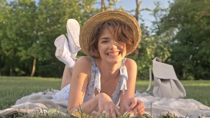 Wall Mural - Cute young woman reading book and smiling looking at the camera while lying on a plaid in the park 