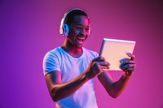 Young african-american man's listening to music in headphones, using tablet in neon light on gradient background. Concept of human emotions, facial expression, summer holidays or weekend, hobby,