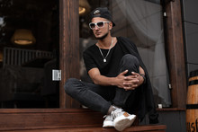 Young Hipster Male In Black Trendy Cap In Fashionable Sunglasses In A T-shirt In Jeans In Sneakers Sits On A Wooden Vintage Staircase Near A Cafe In The City. Attractive Stylish Guy Relaxing Outdoors.