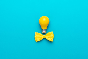 Top view photo of smart idea with yellow bulb and bow tie. Minimalist flat lay image of bow tie and lightbulb over turquoise blue background with copy space. Central composition of great idea concept.