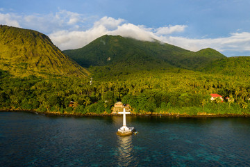 Wall Mural - Aerial drone view of a large cross marking a sunken cemetery with evening light on background volcanos on a tropical island (Camiguin, Philippines)