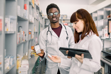 Wall Mural - Two pharmacist looking for medicinal drug. Male African pharmacist holding a medicine with female standing by and using digital tablet in pharmacy.