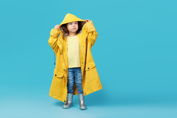 Happy funny child posing on blue studio background. Girl is wearing yellow waterproof raincoat and rubber boots. Weather forecast concept