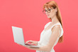 © LIGHTFIELD STUDIOS - serious redhead young woman in glasses using laptop isolated on pink