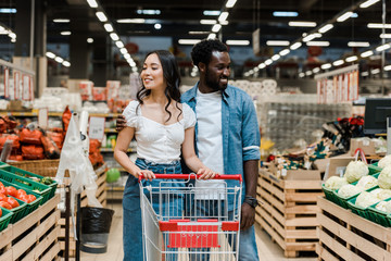 cheerful african american man and happy asian woman near shopping cart in supermarket