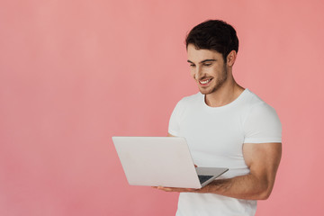 Wall Mural - smiling muscular man in white t-shirt using laptop isolated on pink