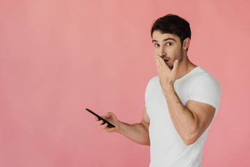 Wall Mural - surprised muscular man in white t-shirt using smartphone and covering mouth with hand isolated on pink