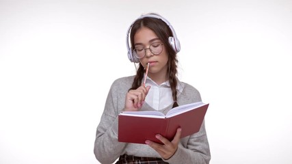 Canvas Print - Concentrated young brunette girl student smiling and writing in notebook while listening an audio book with wireless headphones over white background isolated