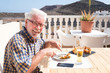 © luciano - Happy adult senior man have a break on a wooden table looking at mobile phone. One people only. Vegetarian food.Mountain on background