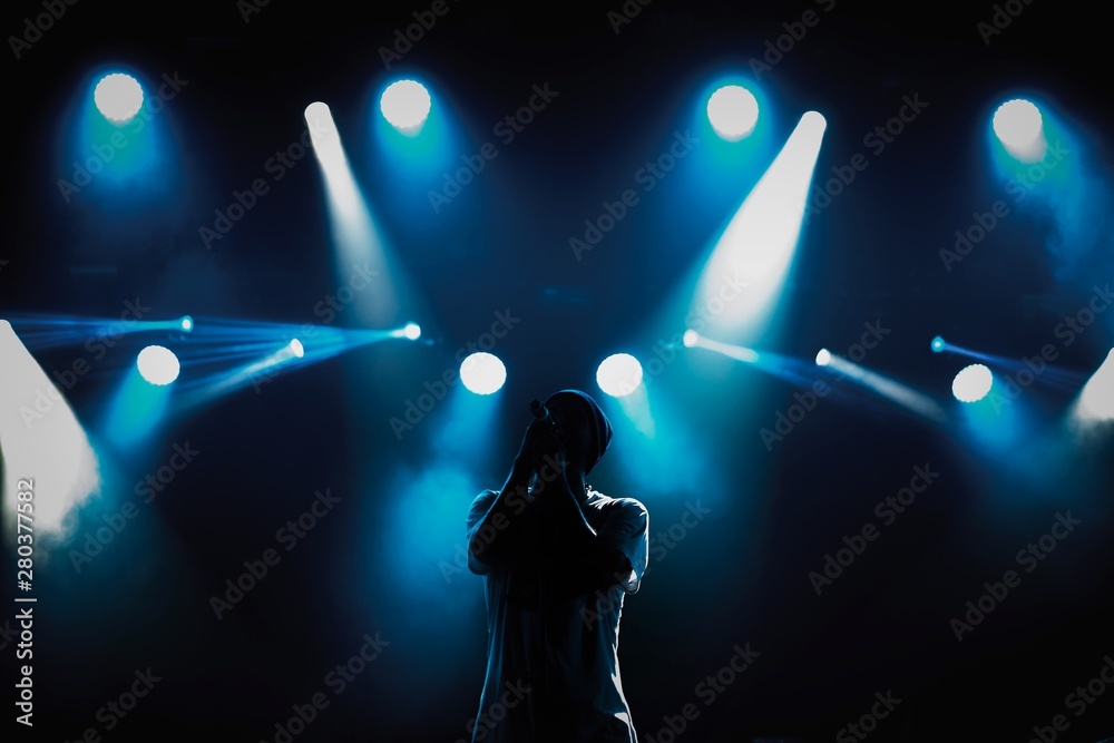Rap singer with microphone on stage in music hall.Silhouette of hip hop singer with mic in hands singing a song on scene in blue lights.Young pop musician sings popular song on stage in music hall - obrazy, fototapety, plakaty 
