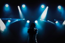 Rap Singer With Microphone On Stage In Music Hall.Silhouette Of Hip Hop Singer With Mic In Hands Singing A Song On Scene In Blue Lights.Young Pop Musician Sings Popular Song On Stage In Music Hall