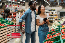 Cheerful African American Man Near Attractive Asian Woman Holding Notebook In Supermarket