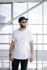 Wall Mural - Hipster handsome male model with beard wearing white blank t-shirt and a baseball cap with space for your logo or design in casual urban style