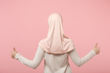 Back rear view of young arabian muslim woman in hijab light clothes posing isolated on pink background studio portrait. People religious Islam lifestyle concept. Mock up copy space. Showing thumbs up.