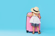 © oes - Happy child girl tourist packs clothes into a suitcase for travel, summer vacation. Toddler kid in straw hat  holding a pink suitcase on a blue background. View from the back