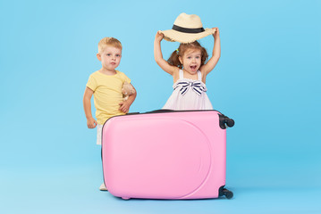 Wall Mural - Happy kids sitting on colorful pink suitcase prepared for summer vacation. Young travelers. Little girl and boy, sister and brother, having fun isolated on blue background