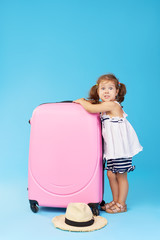 Wall Mural - Happy child girl tourist packs clothes into a suitcase for travel, summer vacation. Toddler kid in straw hat  holding a pink suitcase on a blue background