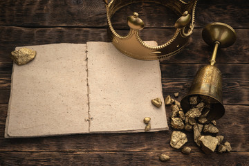 Open blank page book, golden crown and a goblet full of gold on a wooden table background.