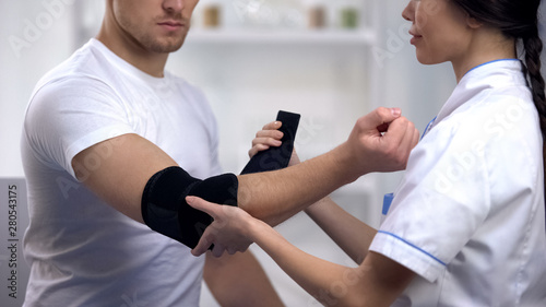 Cuadros en Lienzo Smiling doctor fixing elbow padded orthosis to sportsman patient, rehabilitation