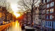 Sunset Reflected In The Water Of The Amsterdam Canal Against The Background Of Beautiful Houses, Trees With Parked Boats, Bicycles And Cars With The Silhouette Of A Church In The Distance, Time Lapse