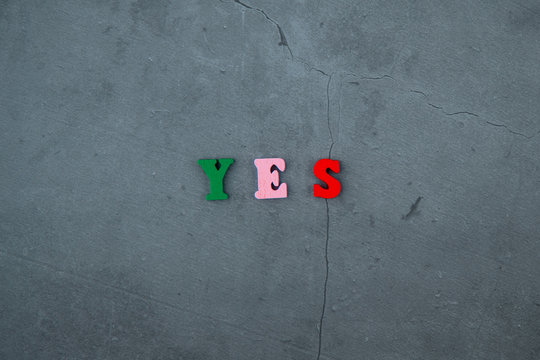 The multicolored Yes word is made of wooden letters on a grey plastered wall background.
