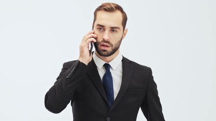 Poster - Serious young businessman in black suit and blue tie becoming angry and gesturing with hand while having a call over gray background isolated