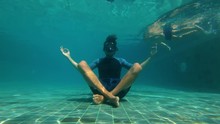 Slowmotion Underwater Shot Of A Young Man In A Yoga Pose Meditating On The Bottom Of A Swimming Pool. He Is Disturbed By A Little Boy Swimming Nearby. Concept Of Impossibility Of Complete Detachment