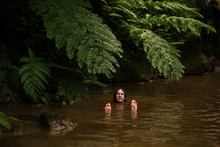 A Curly Man Swimming On His Back On The Silty Brown Water With Face And Feet Raised Up. Tropical River Covered With Big Fern Leaves
