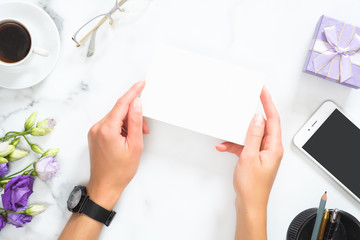 Female hands holding blank paper card mockup over modern home office desk workspace with stationery, cup of coffee, flowers bouquet, glasses, smartphone. Flat lay, top view