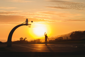 Male plays basketball alone while the sunsets behind him outdoors