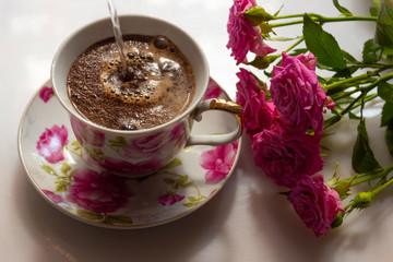 Hot water is poured into a cup for making morning coffee. A cup of coffee with pink decor and flowers of roses on the table in the morning sun.