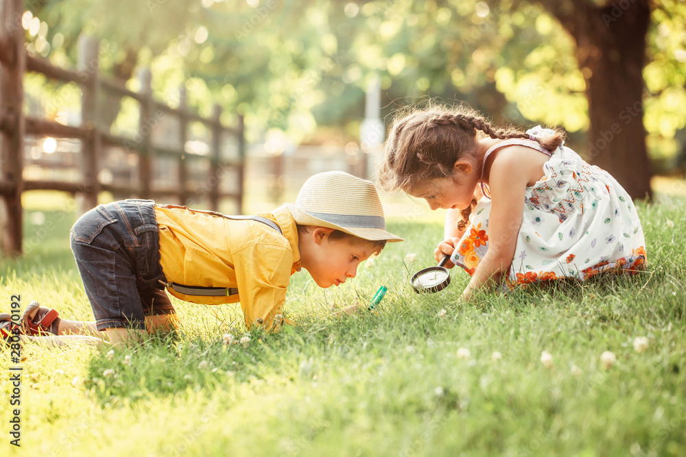 Cute adorable Caucasian girl and boy looking at plants grass in park through magnifying glass. Children friends siblings with loupe studying learning nature outside. Child education concept. - obrazy, fototapety, plakaty 