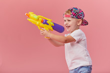 Happy Little Toddler Boy  In Summer Clothes Holds Toy Water Gun Isolated On Pink Wall Background. Children Studio Portrait. People Childhood Lifestyle Concept.