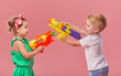 © oes - Happy little toddler boy and girl  in summer clothes holds toy water guns isolated on pink wall background. Children studio portrait. People childhood lifestyle concept.