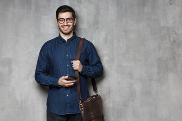 Wall Mural - Modern entrepreneur standing against gray textured wall with copy space, holding phone in hand and bag on shoulder