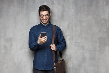 Wall Mural - Modern smiling entrepreneur in glasses and denim shirt standing against gray textured wall with smart phone in hand and bag on shoulder