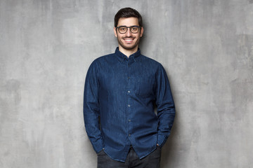 Wall Mural - Young handsome man wering eyeglasses, smiling and feeling confident, standing against gray textured wall background